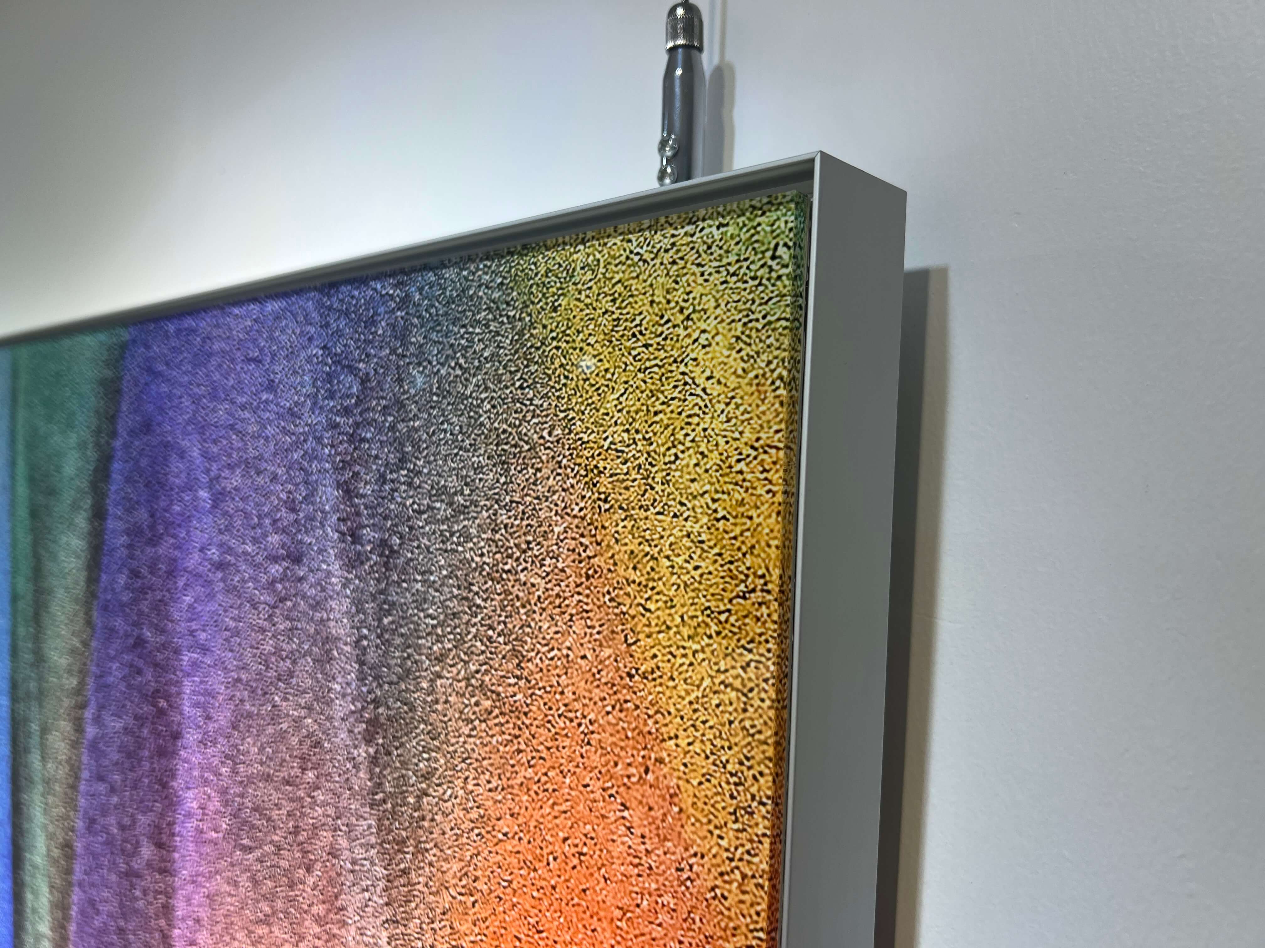 This photo shows a corner of a framed artwork of 'Emotional Spectrum'. You can see the edge where the colours meet the simple, silver frame.  The artwork is hung on a white wall, and there's a small shadow behind the frame. It's like a little piece of a rainbow caught inside a frame on the wall.