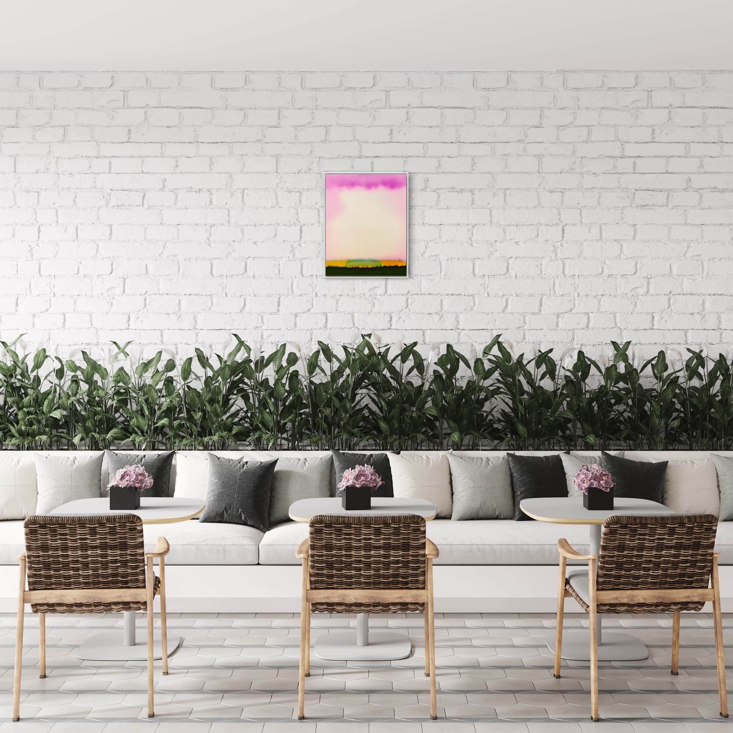 The image shows a bright, airy room with a white brick wall where "Heavy Cloud I" is hung, a small artwork with a prominent pink cloud at the top and a strip of green at the bottom. Below the artwork, there’s a long, continuous gray sofa lined with cushions, in front of which sit two rattan chairs with white tables. Behind the sofa, a row of lush green plants adds a touch of nature to the space. The room’s floor is tiled in a herringbone pattern, contributing to the clean and modern feel of the setting.