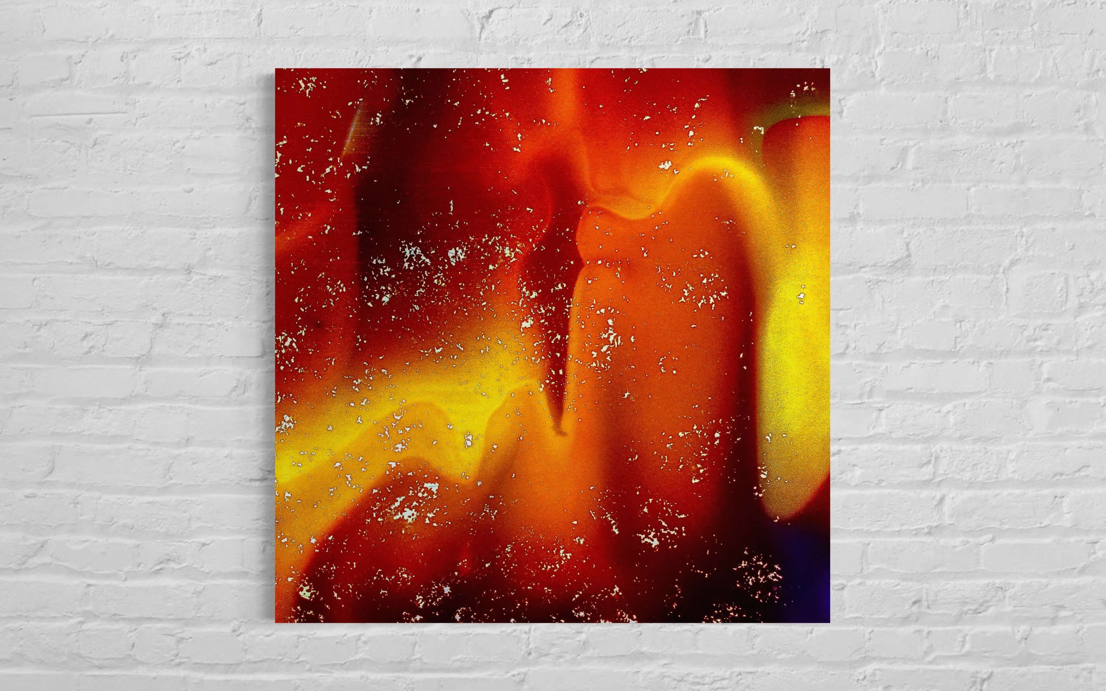 This image shows "Flaming," an artwork full of warm colours like red and yellow, mounted on a white brick wall. The colours blend together and have a fiery look, with little white specks scattered across, like embers in a fire. The artwork stands out against the wall and seems to bring a warm glow to the room.